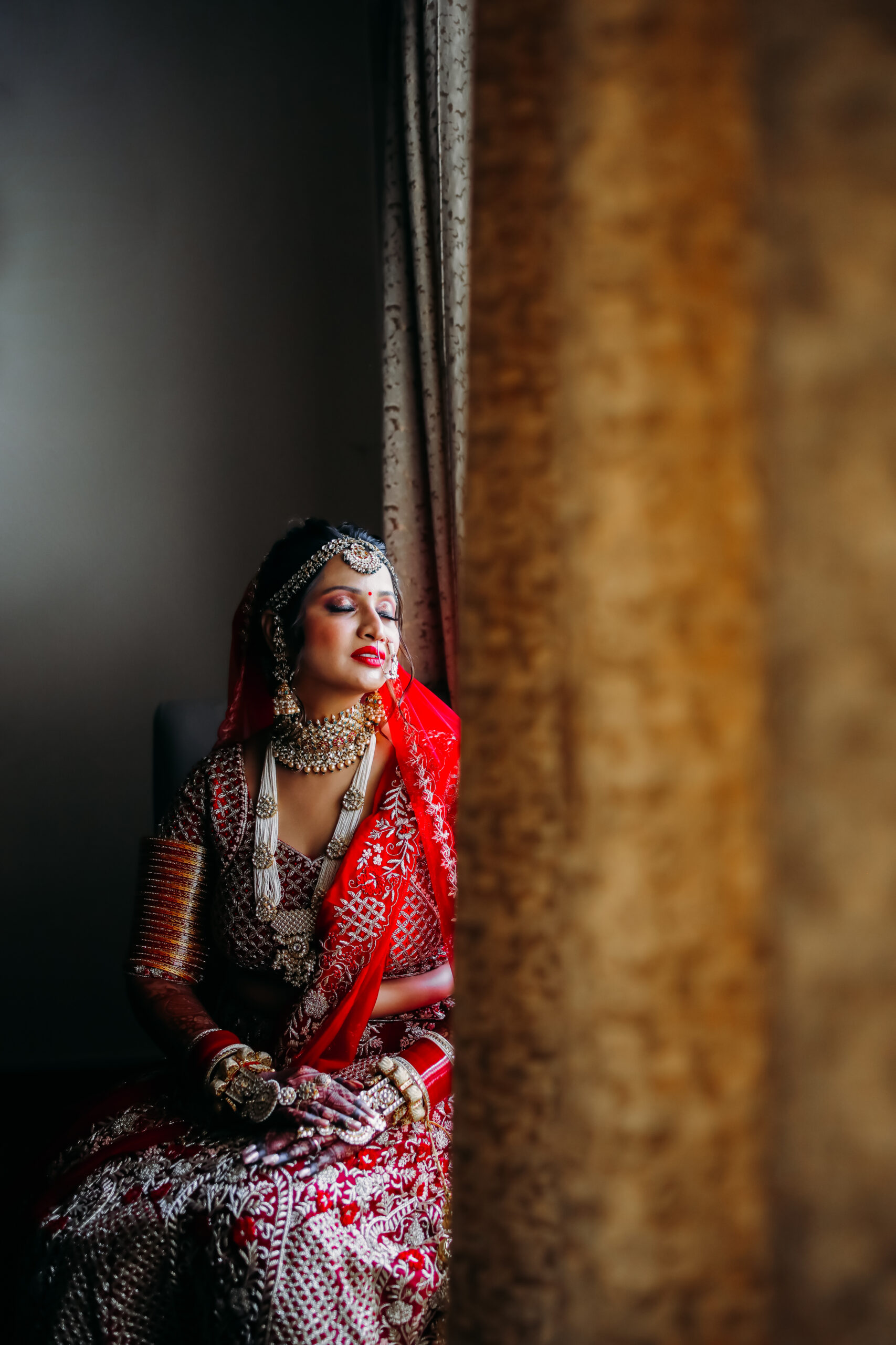 Indian bride in a red embroidered lehenga with traditional jewelry, sitting by a window with soft natural light, eyes closed and hands adorned with mehndi, creating a serene and elegant portrait.
